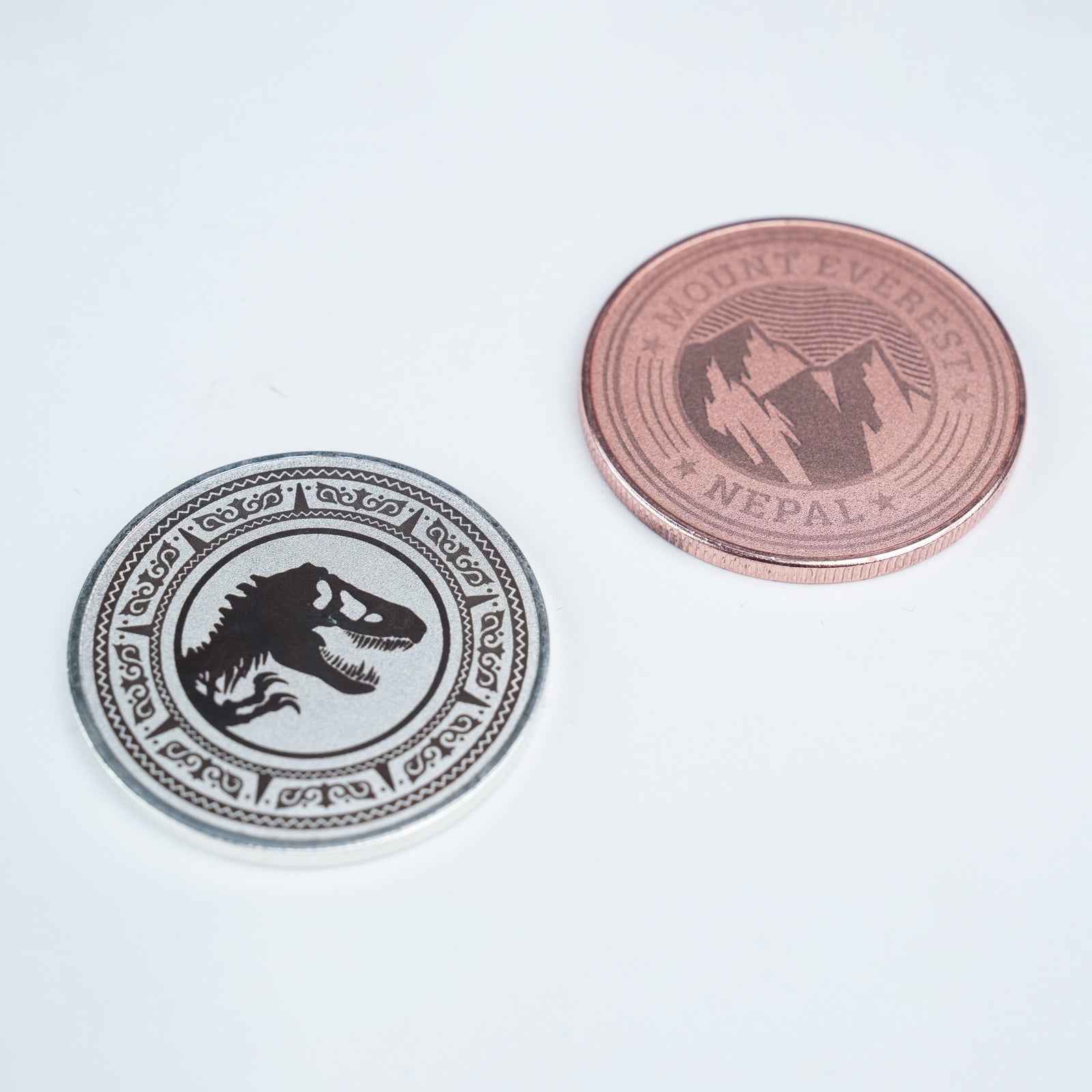 Two Creality Falcon 3-Color Blank Commemorative Coins for Laser Engraver (6pcs) are shown on a white surface: one silver with a dinosaur skull design, and one copper featuring mountains and "Mount Everest Nepal" text.